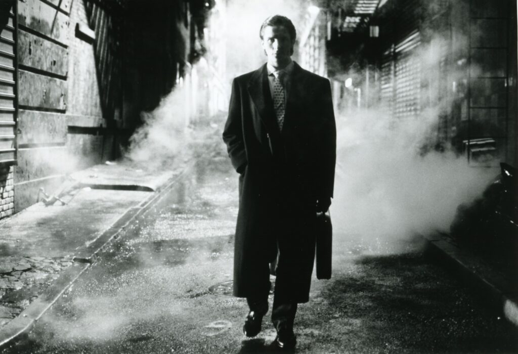 一张黑白照片，一个男人(克里斯蒂安·贝尔饰)穿着黑色西装和黑色长外套，拿着一个公文包。烟雾缭绕，他走在一条黑暗的小巷里。