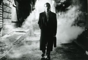 一个男人(克里斯蒂安·贝尔)的黑白照片，穿着黑色西装和黑色长外套，拿着一个公文包。他走在一条黑暗的小巷里，烟雾包围着他。