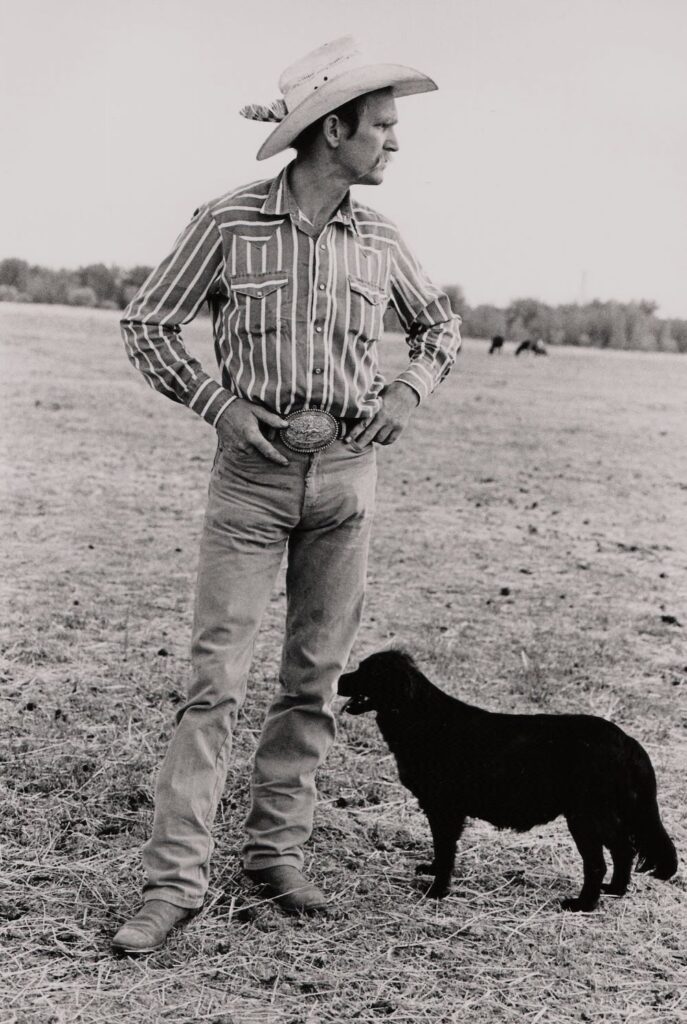 一名男子戴着带有特色的浅色牛仔帽，身穿条纹西式衬衫、牛仔裤和靴子，站在泥泞和干燥的草地上，身边有一只中等大小的深色狗。