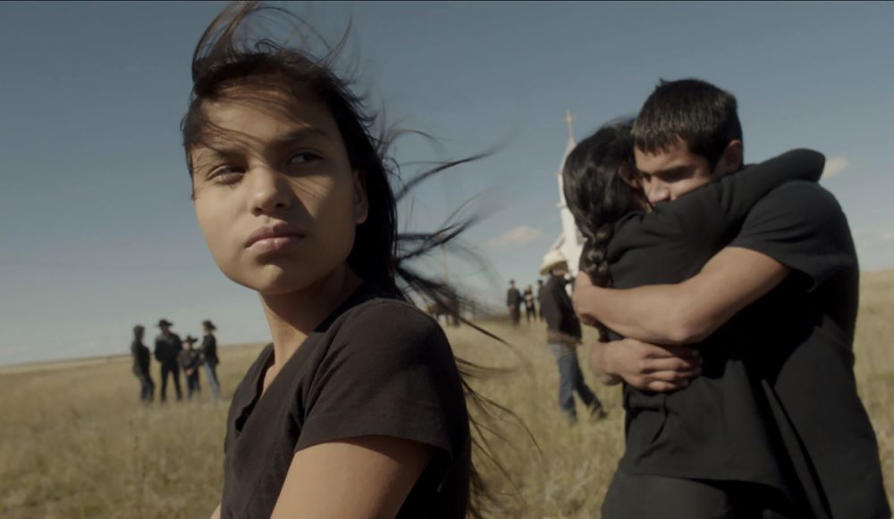 一个年轻的土著女孩站在草原上，近背景中是一对年轻的男女拥抱，背景中是穿着西式服装的母亲和一座教堂