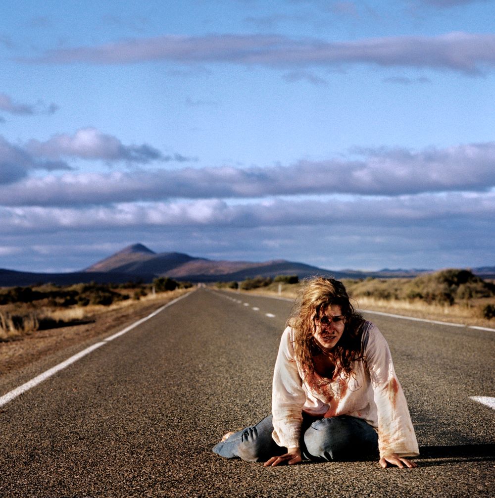 在空旷的公路中央，一个头发缠结的、鲜血淋漓的女人坐着，双腿交叉在身下，背景是荒凉的山脉。