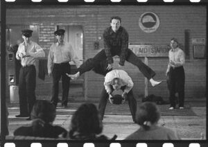在一个临时搭建的舞台上，一名男子跳过另一名男子，身后还有另外三名男子，其中两名戴着警察帽