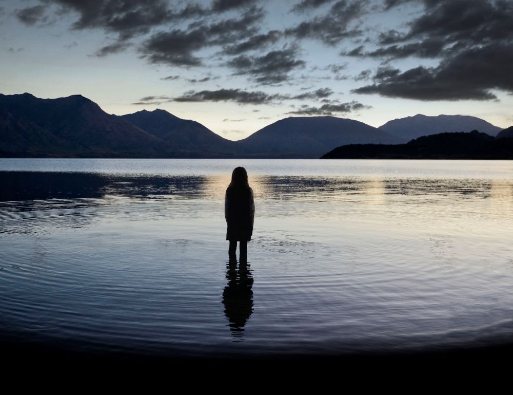 在群山和多云的夜空的背景下，一个女孩站在水中，背朝着镜头。