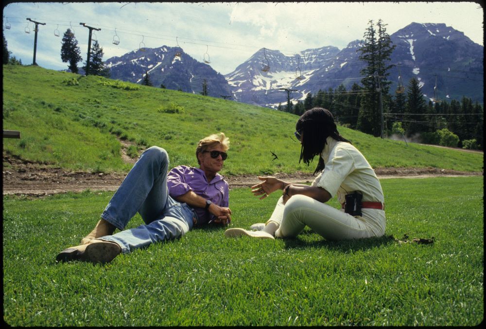 在绿色领域在山地环境中,在后台升降椅,一个blone男人戴着墨镜,牛仔裤,浅紫色拍摄靠背在白人对黑人妇女slcks和衬衫,盘腿坐着,脸离相机