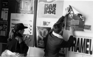 一名男子背对着镜头，在挂满海报的墙上挂着一张写着“瓶子火箭”的海报。一个棕色长发扎着马尾，头戴棒球帽的女人，手里拿着一卷胶带。