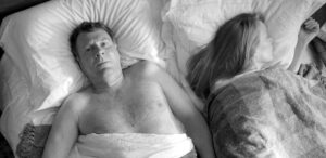 一对中年夫妇躺在床上,赤裸上身的男人躺在他的背上,盯着天花板。在右边,与浅棕色的长发女人躺在她的身边,似乎睡着了,面对远离男人,