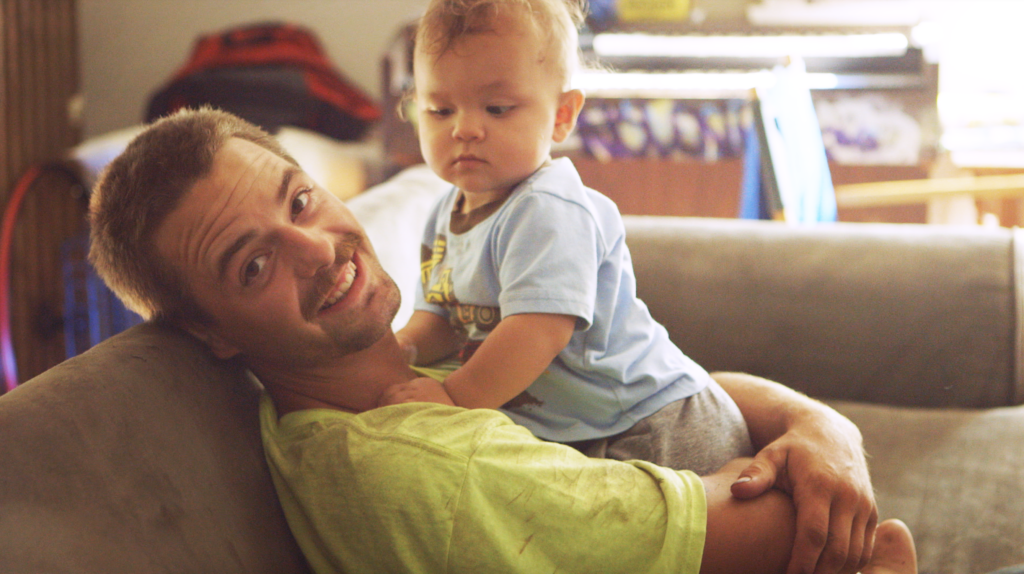 男人在他20多岁的举着一个婴儿坐在沙发上对着镜头微笑