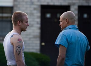 两个男人站在支持主要是相机,面对彼此。左边的男人寸头brown-red头发,纹身在他的手臂和肩膀;他穿着一件白色的无袖汗衫。右边的人似乎有一个光头,身穿蓝色短袖衬衫。