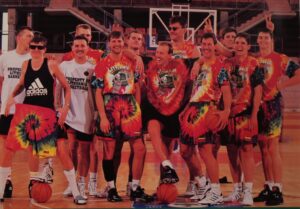 一支兴高采烈的篮球队站在篮球场上合影，挥舞着胜利的手势，许多人穿着或手持彩色扎染篮球短裤