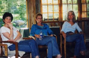 三个女人坐在一个房间里的弧形椅子上，房间里有常青树，窗外有一条小路。所有人都穿着随意的牛仔裤。